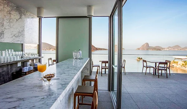 Sala de refeições com vista panorâmica para o mar e montanhas ao fundo, decorada com mesas e cadeiras elegantemente dispostas durante o dia.