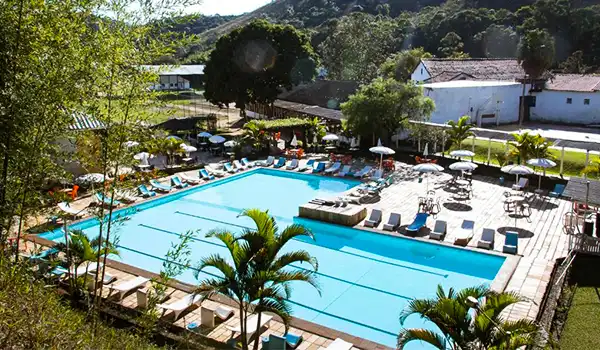 Piscina ao ar livre rodeada de cadeiras de descanso, guarda-sóis e vegetação, em uma área de resort ou hotel com vista para áreas verdes e relevo acidentado ao fundo. Hotéis para Lua de Mel no estado do Rio de Janeiro.