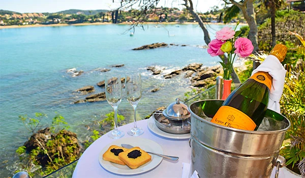 Mesa ao ar livre com champanhe, duas taças, biscoito decorado e flores rosa, ao lado de um lago com árvores e pedras, ideal para momentos românticos ou celebrações ao ar livre. Armação dos Búzios hotéis para Lua de Mel.