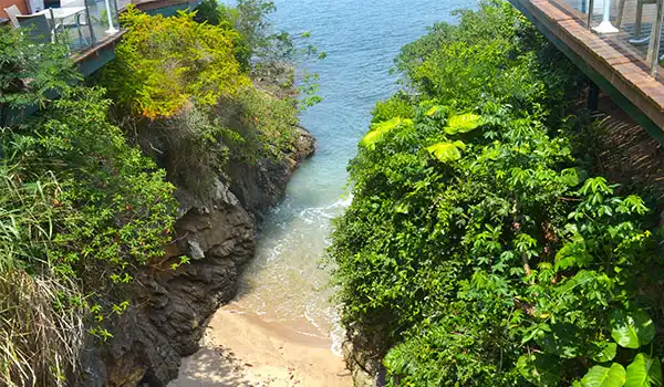 Praia escondida entre rochas e plantas verdes, com vista para o mar em um cenário tropical tranquilo e paradisíaco.