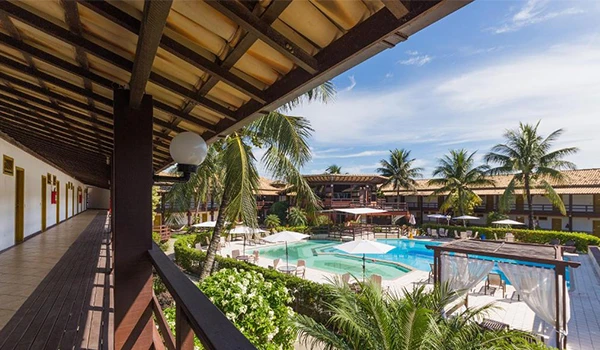 Imagem de uma área de lazer com piscina, cercada por palmeiras e cabanas em um resort tropical, sob céu ensolarado.