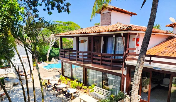 Imagem de um hotel resort com fachada de madeira e piscina ao fundo, rodeado por árvores e plantas tropicais, ideal para férias de lazer.