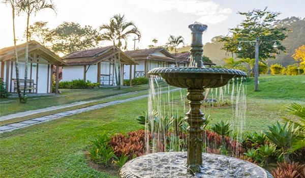Fonte de água em um jardim bem cuidado, com vegetação exuberante, casas de madeira ao fundo e árvores, criando um ambiente relaxante e natural.