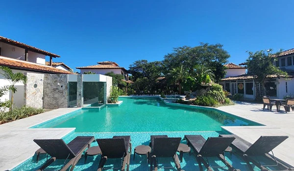 Piscina de luxo ao ar livre com espreguiçadeiras, cercada por casas modernas, árvores verdes e um céu claro, ideal para relaxar e aproveitar dias ensolarados.