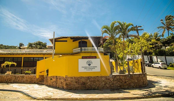 Fachada de Pousada com destaque para sua cor amarela e estrutura moderna, localizada em uma área com árvores e veículos ao redor.