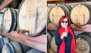 Deusa Rodrigues degustando vinho em adega com barris de madeira marcados 'Três Pedras', cenário de vinícola.