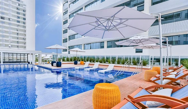 Piscina na cobertura de um prédio moderno com espreguiçadeiras, guarda-sóis brancos e lounge ao ar livre em um dia ensolarado. Hotéis para Lua de Mel no estado do Rio de Janeiro.