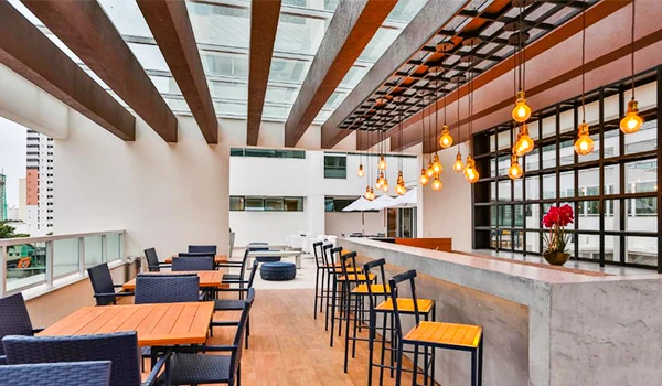 Varanda de restaurante com mesas e cadeiras ao ar livre, iluminação aconchegante com pendentes âmbar, vista moderna de edifícios ao redor, ideal para refeições ao ar livre.
