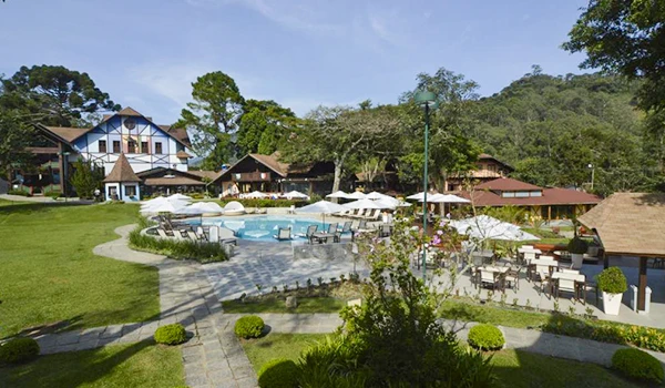Imagem de um resort com piscina ao ar livre, área de lazer com espreguiçadeiras, cadeiras e mesas sob guarda-sóis, rodeado por árvores verdes e edifícios aconchegantes, ideal para relaxar e aproveitar dias de descanso em meio à natureza brasileira. Hotéis para Lua de Mel no estado do Rio de Janeiro.