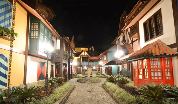 Imagem de um centro histórico com construções coloridas e iluminação noturna, destacando a arquitetura colonial e uma praça central bem cuidada.