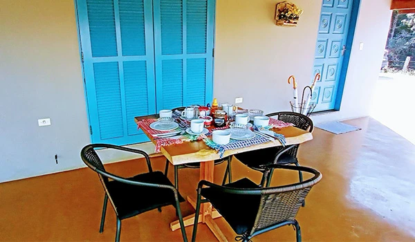 Mesa de jantar ao ar livre, decorada para uma refeição ao ar livre com pratos, copos e utensílios, em um ambiente com portas azuis ao fundo.