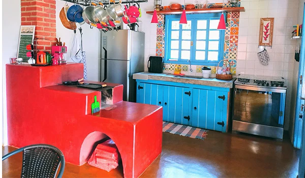 Cozinha colorida com armário azul, parede de tijolos vermelhos, utensílios pendurados e uma janela com cortina azul.