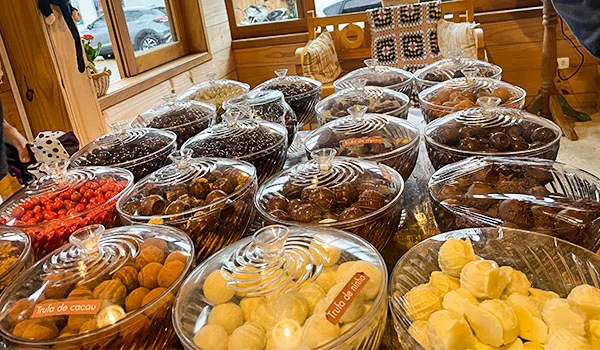 “Vários doces artesanais de diferentes cores e formatos estão dispostos em recipientes transparentes com tampas sobre uma mesa de madeira. Alguns recipientes têm etiquetas como ‘Trufa de cacau’ e ‘Trufa de limão’. Ao fundo, há janelas de madeira e uma decoração aconchegante.”