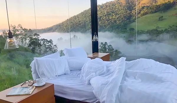 Cama de casal em um quarto com vista panorâmica de uma paisagem natural, incluindo montanhas, árvores e neblina ao amanhecer, criando um ambiente relaxante. Chalés em São Francisco Xavier.