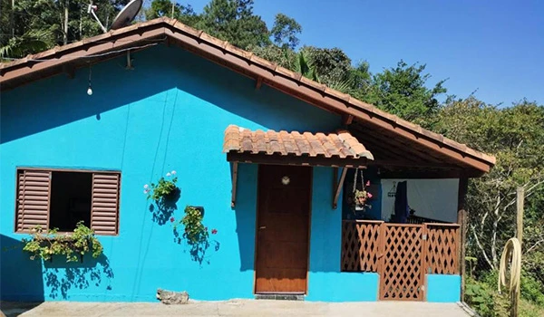 Casa de cor azul claros com varanda de madeira e jardim em um ambiente natural, cercada por árvores verdes sob céu claro.