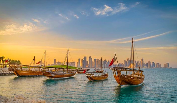Quatro barcos de madeira ancorados na lagoa de Doha, com vista para o horizonte de arranha-céus ao entardecer, refletindo uma cena tranquila e moderna.