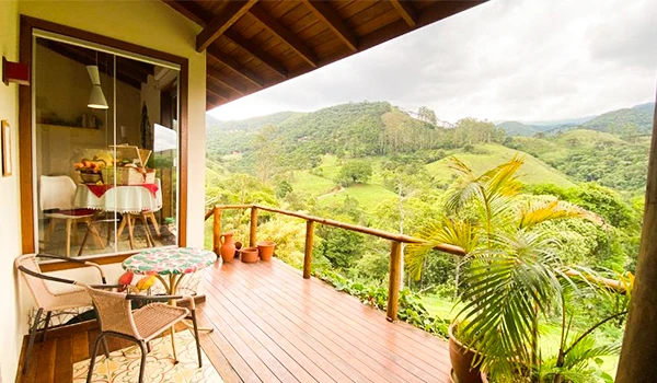 Varanda com vista para a natureza exuberante, com cadeiras de fibra, mesa, plantas e uma paisagem de montanhas verdes ao fundo, ideal para relaxar. Chalés em São Francisco Xavier.