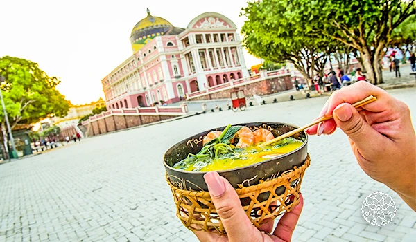 Mulher segurando uma tigela de comida típica ao ar livre em frente ao Palácio Belvedere, em Ubud, Bali, na Indonésia, durante o dia, com árvores verdes ao redor.