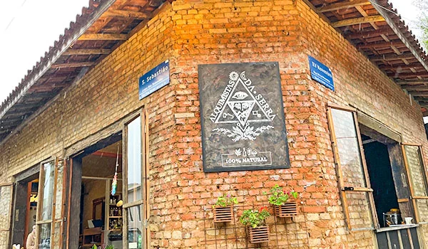 Edificação rústica de tijolos aparentes e telhado de telhas, com portas e janelas abertas. Na parede central, há uma placa com o nome “Alquimista da Serra” e a indicação “100% Natural”. Pequenos vasos com plantas decoram a fachada.