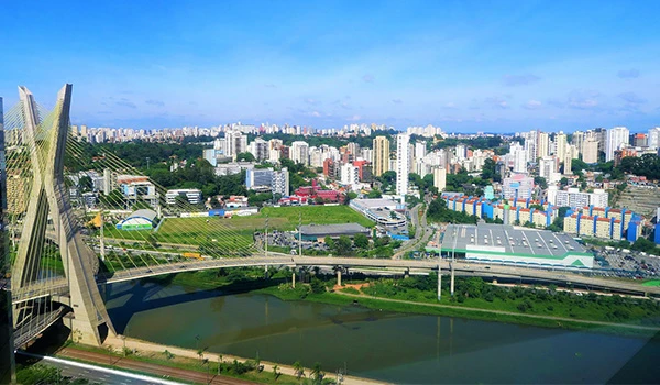 Vista panorâmica da cidade de Curitiba, mostrando a ponte, rios, bairros residenciais e prédios altos sob céu azul claro, destacando o skyline local.