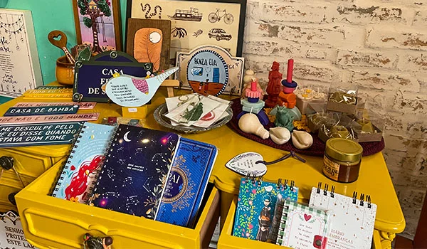 “Balcão amarelo repleto de objetos decorativos e itens artesanais, incluindo cadernos, livros, cartões, pequenos brinquedos, esculturas e potes. Ao fundo, há uma parede branca de tijolos aparentes com quadros e ilustrações, criando um ambiente colorido e descontraído.”
