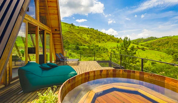 Imagem de uma varanda com vista para uma paisagem verdejante, incluindo uma banheira de hidromassagem de madeira ao ar livre, em um ambiente de montanha. Perfeita para relaxamento e conexão com a natureza, ideal para quem busca tranquilidade e conforto em um refúgio na natureza. Chalés em São Francisco Xavier.