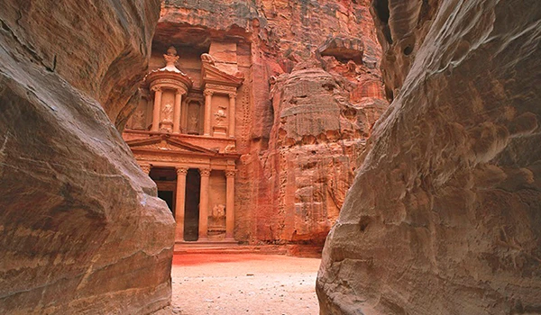 Imagem do Al Deir, antigo templo na cidade de Petra, na Jordânia, construído em pedra rosa, cercado por rochas e formações rochosas impressionantes.