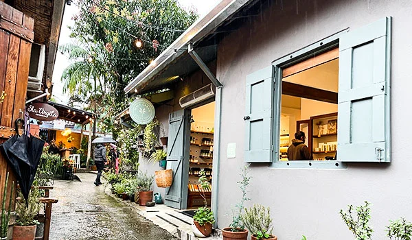 “Pequeno espaço comercial charmoso em uma área arborizada, com paredes claras, janelas e portas de madeira azul-clara abertas, plantas em vasos e iluminação aconchegante. Ao fundo, há um caminho de pedras, mesas ao ar livre e outros estabelecimentos.”