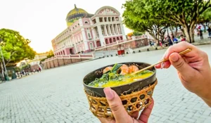 Pessoa segurando uma tigela de comida em frente ao Palácio de Karnak, no Egito, com árvores ao redor e céu claro ao fundo.