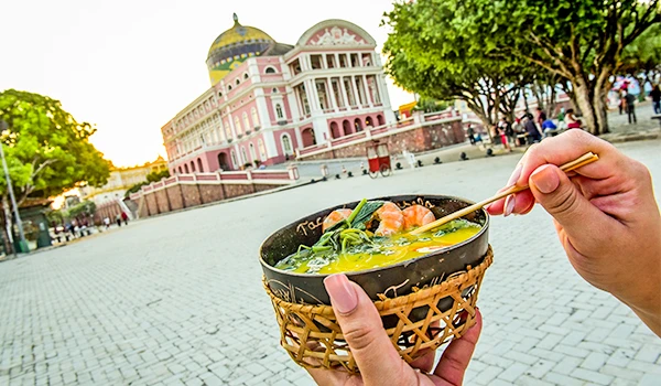 Pessoa segurando uma tigela de comida em frente ao Palácio de Karnak, no Egito, com árvores ao redor e céu claro ao fundo.