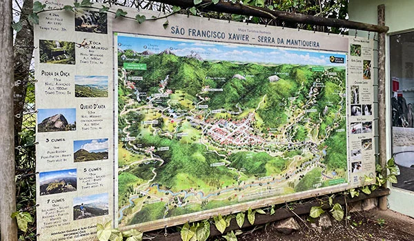 Painel turístico ilustrado com mapa de São Francisco Xavier, na Serra da Mantiqueira, cercado por árvores e vegetação. O mapa destaca trilhas, montanhas, rios, cachoeiras e pontos turísticos da região.