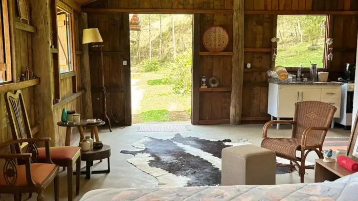 Interior de uma cabana rústica com móveis de madeira, tapete de couro, cadeira de vime e vista para a natureza ao fundo, ideal para relaxar no campo.