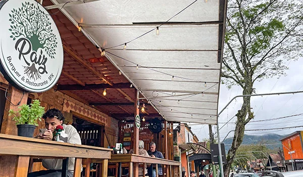 Fachada do restaurante Raiz Burger & Restaurante, com mesas de madeira sob uma cobertura iluminada por cordões de lâmpadas. Algumas pessoas estão sentadas no espaço externo. Ao fundo, aparecem árvores, construções e montanhas da região.