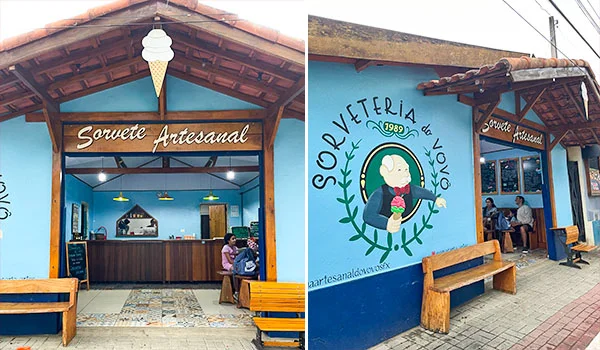 Fachada de uma sorveteria artesanal pintada de azul, com telhado de cerâmica, bancos de madeira e mesas na calçada. À esquerda, a entrada exibe a inscrição ‘Sorvete Artesanal’; à direita, há um mural com o desenho de um personagem segurando um sorvete e o nome ‘Sorveteria do Vovô’.”