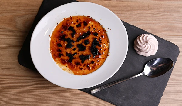 Close-up de uma sobremesa de creme queimado em prato branco, acompanhado de chantilly ao lado, sobre uma tábua de pedra no fundo de madeira, uma das atrações de Coimbra.