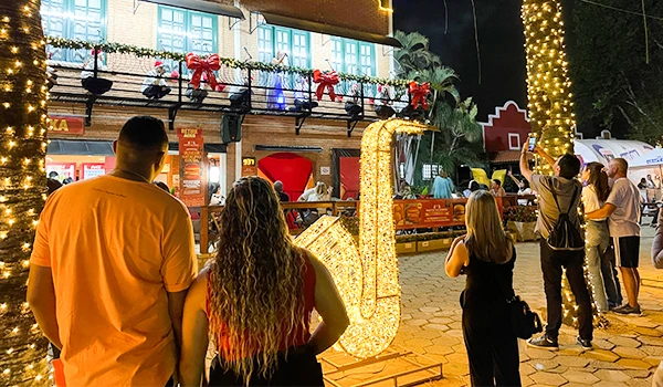 Pessoas apreciando decoração de Natal em uma rua com luzes e um grande enfeite de letra 'L' iluminada, com árvores decoradas e ambiente festivo. Parada de Natal de Holambra.