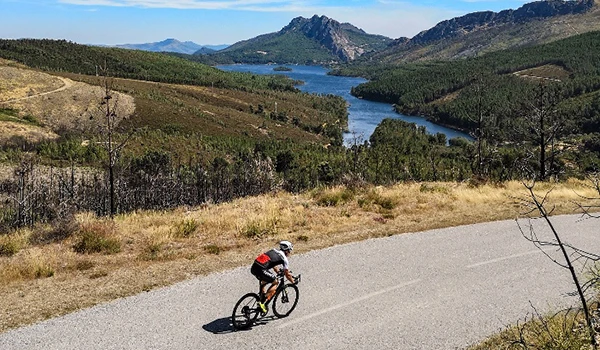 Ciclista pedalando por uma estrada de asfalto em meio a um cenário de montanhas, rio e natureza exuberante, sob céu claro em uma paisagem rural.