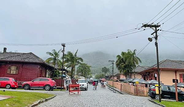 Imagem de uma rua de bairro com calçamento, casas de madeira, árvores e pessoas caminhando sob céu nublado e neblina, com veículos estacionados na rua