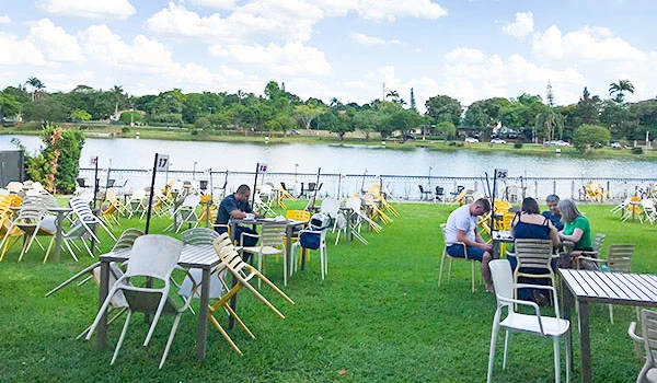 Imagem de um restaurante ao ar livre às margens de um rio, com várias mesas e cadeiras, algumas pessoas desfrutando de refeições, sob céu parcialmente nublado, com árvores ao fundo.