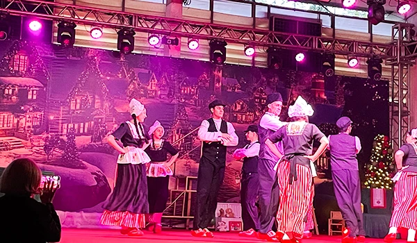 Apresentação teatral com atores vestidos com roupas tradicionais em palco decorado com cenário de cidade antiga, iluminação rosa e pessoas assistindo ao espetáculo. Parada de Natal de Holambra.