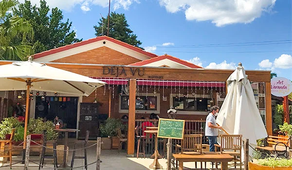 Imagem de uma cafeteria ao ar livre com mesas, cadeiras e sombrinhas, ambiente acolhedor e decorado com luzes de natal, sob céu azul com nuvens brancas.