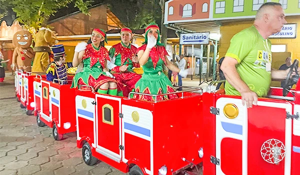 Família participando de passeio natalino com trenó decorado na rua, incluindo pessoas vestidas de Duendes de Papai Noel, durante evento natalino ao ar livre em cidade animada.