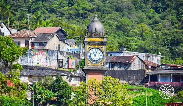 Relógio antigo na praça de uma cidade com casas e árvores ao fundo, em um ambiente de natureza e urbanismo.