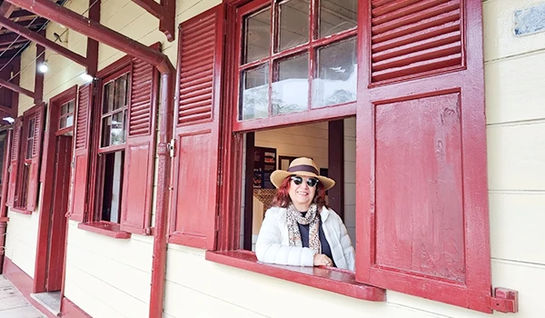 Deusa Rodrigues desfrutando de um dia ensolarado ao lado de uma casa com janelas decoradas com persianas vermelhas.