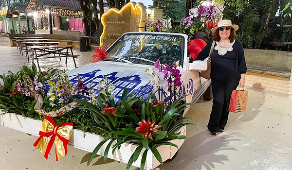 Deusa Rodrigues em evento decorado com flores e carro temático, com pé direito alto e árvore ao fundo. Parada de Natal de Holambra.