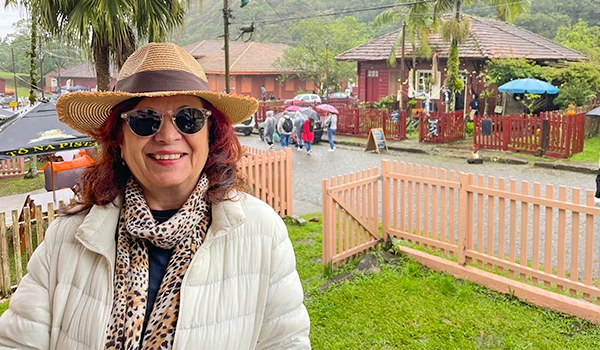 Deusa Rodrigues, óculos escuros e jaqueta bege, passeando sob chuva com guarda-chuva colorido em ambiente ao ar livre, cercada por casas de madeira e árvores verdes.