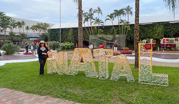 Deusa Rodrigues ao lado de letras decorativas que formam NATAL, em um parque com árvores e pessoas ao fundo. Parada de Natal de Holambra.