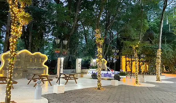 Imagem de uma praça decorada com luzes de Natal em árvores, com bancos, iluminação e uma área de convivência ao ar livre, ideal para momentos de lazer.