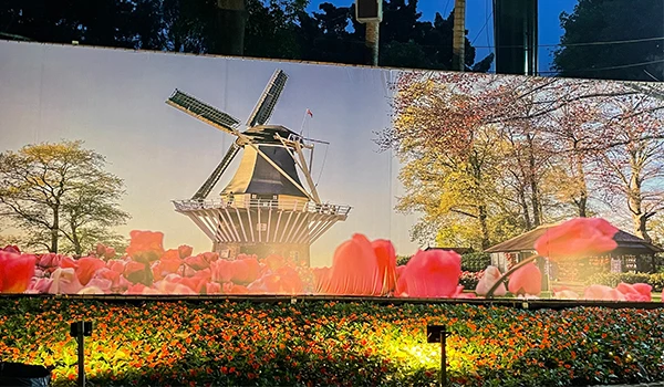 Imagem de uma praça com um moinho de vento tradicional ao fundo, cercada por flores rosa e árvores coloridas, proporcionando um cenário encantador e tranquilo.