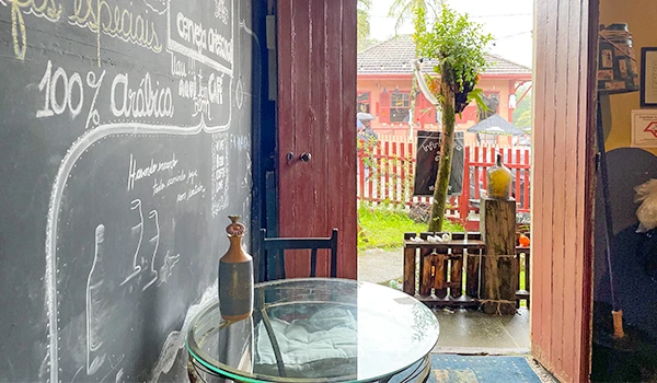 Interior de uma cafeteria com parede de quadro negro, mesa de vidro, e porta que leva a um espaço ao ar livre com árvores e decoração rústica.